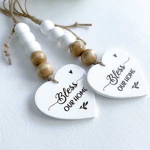 May include: Two white wooden heart-shaped ornaments with the text "Bless Our Home" in brown lettering. Each ornament is attached to a string of white and brown wooden beads.
