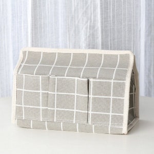 May include: A gray and white checkered tissue box cover with three pockets on the front. The cover is made of fabric and has a white trim around the edges.