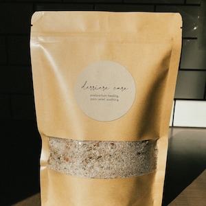 May include: A brown paper bag with a white circle label that says "derriere care postpartum healing, pain relief, soothing". The bag is filled with a white powder.
