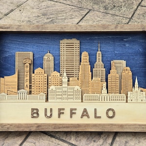 May include: Wooden framed artwork featuring a detailed cityscape silhouette. The artwork showcases a layered design with buildings in varying shades of brown against a blue background. The word "BUFFALO" is spelled out in wooden letters at the bottom.