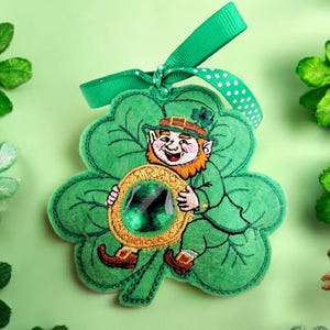 Puede incluir: Adorno de trébol verde de fieltro con un duende sonriente sosteniendo una moneda con borde dorado y gemas verdes. Tiene una cinta verde con lunares blancos para colgar. Una decoración festiva del Día de San Patricio.