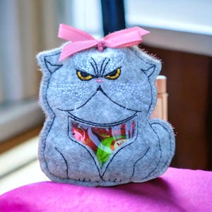 Op de afbeelding: Een grijze vilten katvormige zak met een roze strik erop. De kat heeft geborduurde details, waaronder gele ogen en een humeurige uitdrukking. Een doorzichtige zak in het midden onthult kleurrijke snoepjes. De zak ligt op een roze ondergrond.