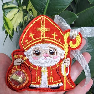May include: Embroidered ornament of a cartoon St. Nicholas figure in a red robe and hat, holding a golden staff and a bag of gifts. The ornament has a silver ribbon for hanging and is set against a background of green leaves.