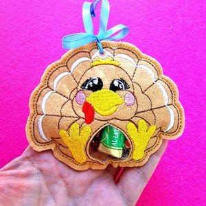 May include: A brown felt turkey ornament with a yellow beak, red wattle, and black eyes. The turkey has a small opening in the back to hold treats or small gifts. The ornament is tied with a blue ribbon.