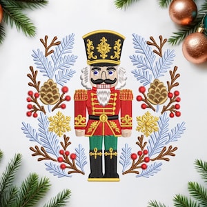 Puede incluir: Diseño bordado de cascanueces navideño en rojo, negro, dorado y verde. El cascanueces está rodeado por una corona de ramas azules y marrones, piñas y bayas rojas. Decoración festiva navideña.