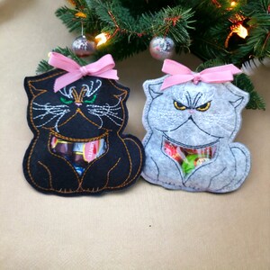 Puede incluir: Dos bolsas de regalo de fieltro con forma de gato con lazos rosas. El gato negro tiene ojos verdes y bigote blanco. El gato gris tiene ojos amarillos y bigote blanco. Ambos gatos tienen una ventana de plástico transparente que muestra caramelos en el interior.