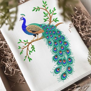 May include: White embroidered towel with a peacock design. The peacock is depicted with blue, green, and orange plumage, perched on a brown branch with green leaves. The towel is folded and presented in a brown box.