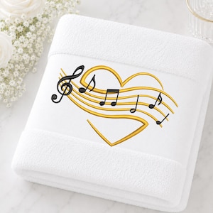 Könnte beinhalten: Ein weißes Handtuch mit einer Stickerei in Form eines goldenen Herzens, das aus Notenlinien besteht. Schwarze Musiknoten und ein Violinschlüssel sind auf den goldenen Linien angeordnet und bilden ein detailliertes, strukturiertes Motiv.