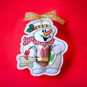 Op de afbeelding: Een witte vilt sneeuwman ornament met een rood-wit geruite sjaal, die een doorzichtige plastic zak met chocolade snoepjes vasthoudt. De sneeuwman heeft een zwarte geborduurde glimlach, zwarte ogen en een oranje wortelneus. Het ornament heeft een gouden glitter lint aan de bovenkant.