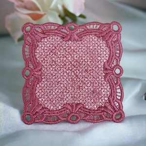 Könnte beinhalten: Eine quadratische, rosafarbene Spitzenunterlage mit einem zarten, filigranen Design. Die Unterlage hat einen gewellten Rand und eine leicht erhabene Mitte.