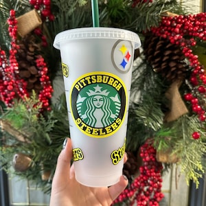 May include: A white reusable cup with a black and yellow Pittsburgh Steelers logo. The logo features a green and white Starbucks logo inside of it. The cup has a black and yellow straw.
