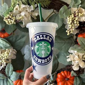 May include: A white reusable cup with a blue and green Dallas Cowboys logo. The cup has a clear lid and a green straw.