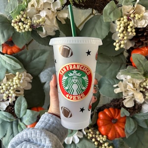May include: A clear plastic Starbucks tumbler with a green straw. The tumbler features a San Francisco 49ers logo, footballs, and black stars. The cup is held in front of a fall wreath with pumpkins and white flowers.