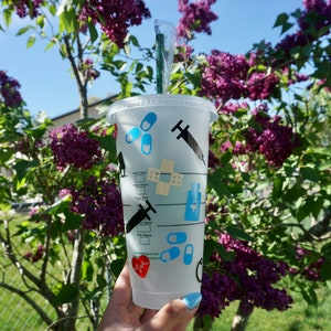 May include: A clear plastic reusable cup with a black and blue medical themed design. The cup has a black straw and is decorated with images of pills, bandages, syringes, and a heart with a pulse line. The cup is held in a hand against a background of a lilac bush.