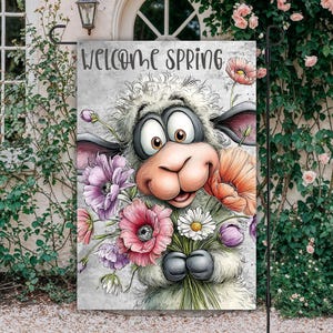 May include: A garden flag featuring a cartoon sheep holding a bouquet of colorful flowers. The flag has the words "WELCOME SPRING" at the top. The sheep has big eyes and a smiling face. The flowers are pink, purple, orange, and white.