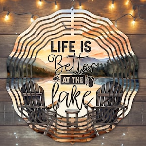 Peut inclure: Un carillon en métal avec une scène de coucher de soleil et le texte "Life is Better at the Lake". Deux chaises sont sur une jetée avec une petite table entre elles.