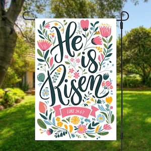May include: A white garden flag with a floral design and the text "He is Risen" in dark blue lettering. A pink ribbon with the text "LUKE 24:6-7" is at the bottom of the flag.