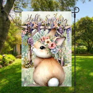 May include: A garden flag with a watercolor illustration of a brown bunny wearing a flower crown. The bunny is standing in front of a wooden trellis with purple flowers. The text "Hello Spring" is written in a whimsical font at the top of the flag.
