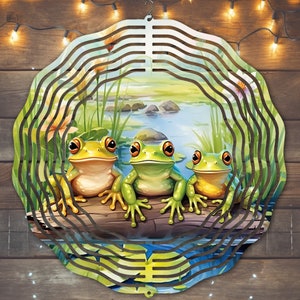 May include: A metal wind spinner with a circular design featuring three green frogs sitting on a log by a pond. The frogs have large, round eyes and are surrounded by green lily pads and reeds.