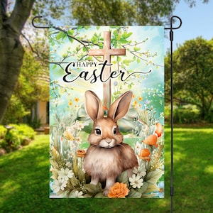 May include: A garden flag featuring a watercolour illustration of a brown bunny rabbit standing in front of a wooden cross. The cross is surrounded by green leaves and flowers. The text "Happy Easter" is written in cursive above the cross.