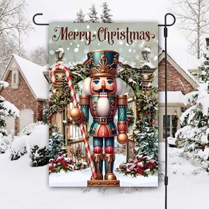 Puede incluir: Una bandera de jardín decorativa con un colorido cascanueces sosteniendo un bastón de caramelo. La bandera dice "Merry Christmas" en una elegante escritura, ambientada en una escena invernal nevada con una casa y árboles cubiertos de nieve.