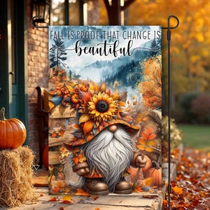 Puede incluir: Una bandera de jardín con un gnomo caprichoso con una larga barba blanca y un sombrero de girasol. La bandera dice "FALL IS PROOF THAT CHANGE IS beautiful" sobre un fondo de follaje otoñal y una escena de montaña.