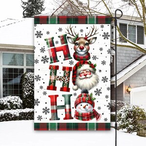 May include: A Christmas garden flag with a white background and the word "HO HO HO" in red, green, and leopard print. The flag features a reindeer, Santa Claus, and a snowman, all wearing festive attire. Snowflakes are scattered around the design.
