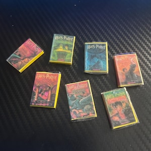 May include: A collection of seven miniature Harry Potter books, each featuring distinct colorful cover art and the Harry Potter title text. The small rectangular books are arranged in a scattered layout, showcasing various illustrated scenes from the series.