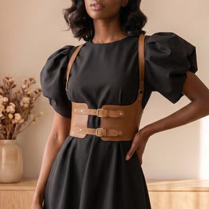 May include: A black midi dress with short puff sleeves and a round neckline, featuring a tan leather-style corset harness worn over the bodice. The harness includes two horizontal straps with metal buckles and vertical shoulder straps.