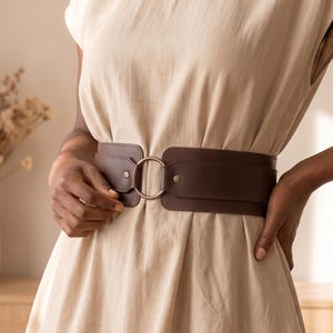 May include: A wide dark brown belt featuring a central circular metal ring buckle. The belt is worn over a light beige sleeveless dress with a simple, clean design. The belt is constructed from two wide panels that meet at the front ring.