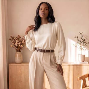 May include: A long-sleeved cream-colored blouse with a round neckline and gathered cuffs, paired with high-waisted beige trousers featuring a front crease and a dark brown leather belt with a silver-toned buckle, creating a coordinated outfit.
