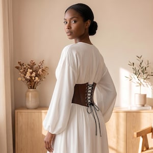 May include: A long-sleeved white dress featuring a brown corset-style waist belt. The belt is secured with black laces that crisscross through metal eyelets and tie in a bow at the center back, creating a cinched silhouette over the fabric.