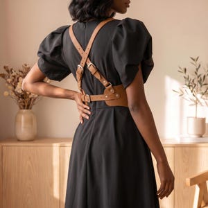 May include: A black dress with short puff sleeves and a tan leather harness featuring adjustable straps, silver-toned metal buckles, and a back-fastening design. The harness is worn over the dress, creating a structured look with visible hardware.