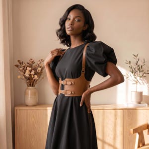 May include: A black dress with short puff sleeves and a round neckline, worn with a tan leather-like corset belt. The belt features two horizontal straps across the front, each secured with a metal buckle, and vertical straps extending upward.