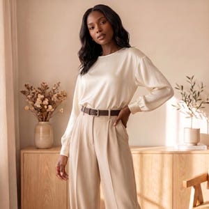 May include: A long-sleeved cream-colored satin blouse with a round neckline and cuffed sleeves, paired with high-waisted beige trousers featuring a dark brown leather belt with a silver-toned buckle. The blouse has a smooth, lustrous finish.