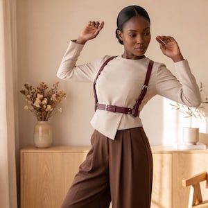 May include: A deep burgundy leather harness belt featuring adjustable straps, silver-toned metal buckles, and a central waist belt. The harness is worn over a long-sleeved beige top and paired with dark brown trousers, creating a structured look.
