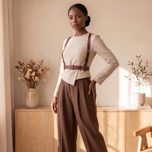 May include: A long-sleeved beige top with a round neckline and an asymmetrical hem, paired with high-waisted brown trousers. A dark burgundy leather harness with silver-toned buckles is worn over the top, featuring a waist belt and shoulder straps.