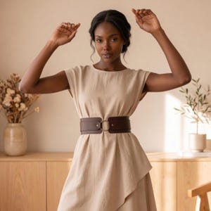 May include: A wide dark brown leather belt featuring a central gold-toned metal ring buckle, worn over a sleeveless beige dress with a round neckline and a draped, asymmetrical hemline that falls below the waist in a soft, flowing silhouette.