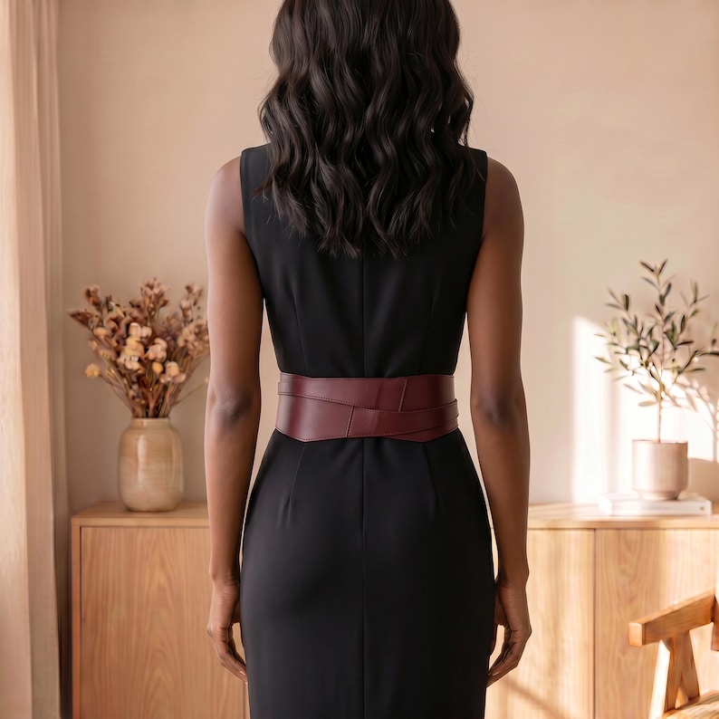 May include: A sleeveless black sheath dress featuring a wide, dark burgundy wrap-style belt cinched at the waist. The dress has a simple, clean silhouette with a fitted bodice and a straight skirt, highlighting the contrast of the deep red belt.