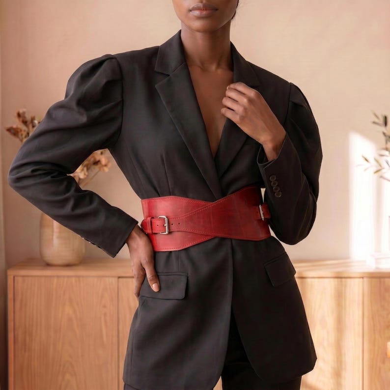 May include: A black blazer with long sleeves, structured shoulders, and a notched lapel, worn with a wide red leather belt featuring a silver-toned metal buckle. The blazer has two front flap pockets and four small buttons on each sleeve cuff.