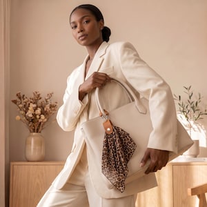 May include: A large, structured beige tote bag featuring two top handles and a detachable tan leather charm. A square, semi-sheer scarf with a brown and black leopard print pattern is tied to the bag's handle with a silver-toned metal clasp.