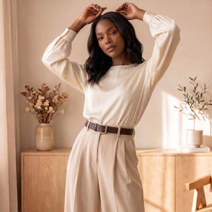 May include: A long-sleeved cream-colored blouse with a smooth, satin-like finish and a round neckline, paired with high-waisted beige trousers featuring front pleats and a dark brown leather belt with a silver-toned rectangular metal buckle.