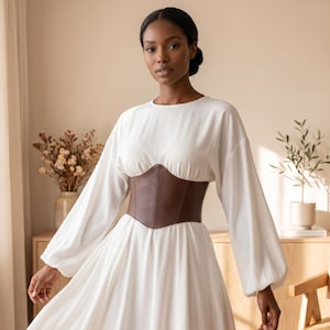 May include: A long-sleeved white dress featuring a round neckline and a structured dark brown faux leather corset belt worn around the waist. The dress has full, flowing sleeves and a simple, clean design that reaches below the waistline.