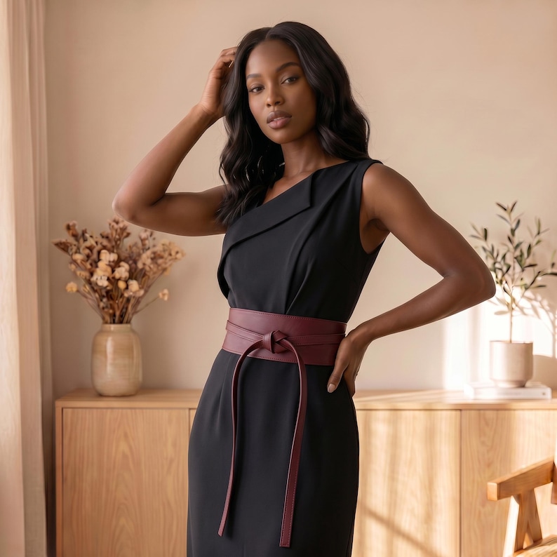 May include: A sleeveless black dress featuring a folded neckline detail and a wide, dark burgundy wrap-style belt that ties at the front with long, hanging ends. The dress has a structured bodice and a clean, solid-colored fabric construction.