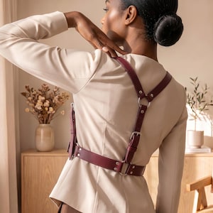 May include: A deep burgundy leather harness worn over a beige blazer. The harness features adjustable straps with silver-toned metal buckles, rivets, and circular rings connecting the back and waist sections in a structured, symmetrical design.
