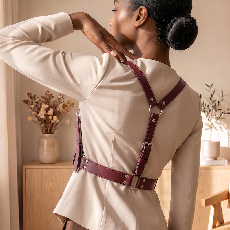 May include: A deep burgundy leather harness worn over a beige blazer. The harness features adjustable straps with silver-toned metal buckles, rivets, and circular rings connecting the back and waist sections in a structured, symmetrical design.