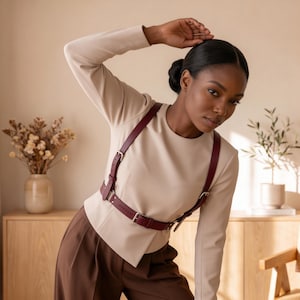 May include: A deep burgundy leather harness featuring adjustable straps and silver-toned metal buckles, worn over a long-sleeved beige top and paired with brown trousers. The harness consists of a waist belt connected to two shoulder straps.