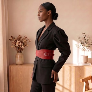 May include: A black blazer with long sleeves and a structured shoulder design, featuring a wide red belt with a silver buckle cinched at the waist. The blazer has a notched lapel, two front flap pockets, and a single-breasted button closure.