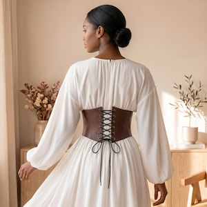 May include: A long-sleeved white dress featuring a brown corset-style waist belt. The belt is constructed with dark brown material, silver-toned metal eyelets, and black crisscross lacing that ties into a bow at the center of the back.
