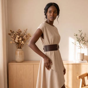 May include: A beige knee-length dress with short sleeves and a round neckline, cinched at the waist with a wide, dark brown leather belt featuring a metallic buckle. The dress has a simple, clean design with a fluid, relaxed fit and hemline.